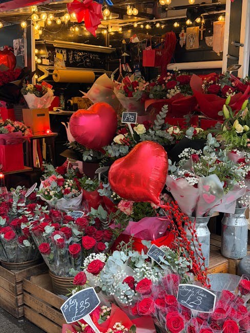 An indoor flower shop display featuring an arrangement of fresh red roses, white baby's breath, and eucalyptus foliage, organized in wooden crates and vases. The display includes red heart-shaped foil