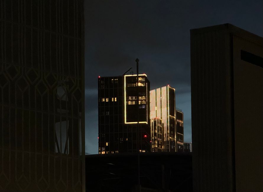 A nighttime view of modern high-rise buildings in London, illuminated with bright white and yellow neon lights, set against a dark, cloudy sky. The buildings have glass facades reflecting the city lig