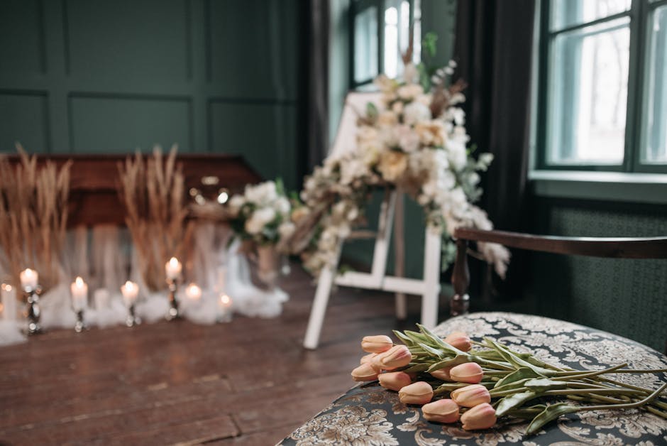 A funeral setting displaying a bouquet of soft pink tulips with green stems resting on an ornate fabric chair in the foreground, with a floral tribute arrangement featuring white roses, lilies, and gr