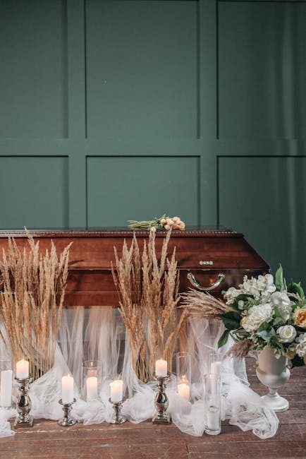 A wooden memorial casket decorated with a small arrangement of pale pink roses and greenery rests on a platform draped with white tulle. In front of the casket, there is a display of tall, dried beige