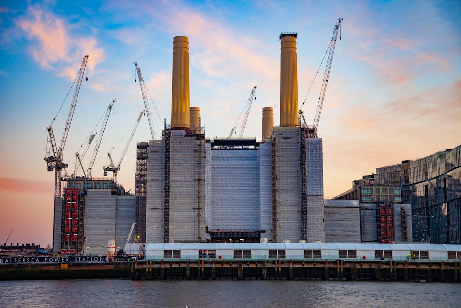 An industrial scene of Battersea Power Station under construction during sunset, with multiple construction cranes surrounding the large, iconic building that features four tall, yellowish chimney sta